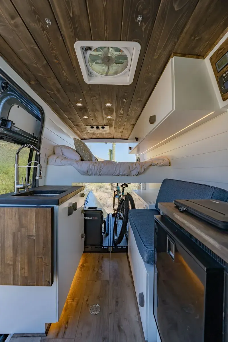 The interior view of the Papago Vans Trail Trekker build. Espresso stained wood with white cabinets and warm ambient lighting under the sink, illuminating the dark-toned wood floors. Fridge, sink, bed lift, and A/C are visible down the middle of the van.