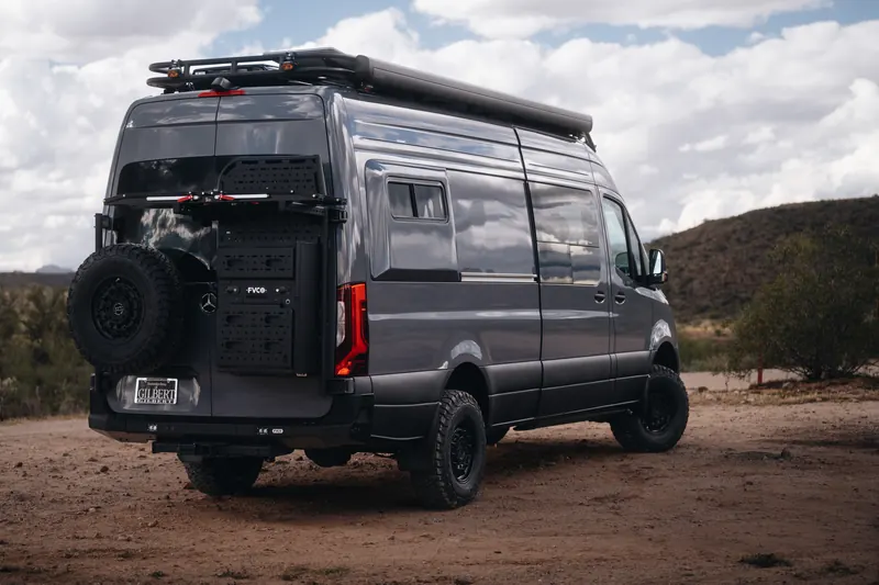 The rear of a grey Papago Sprinter with a spare tire carrier, storage panel and roof rack on a desert road
