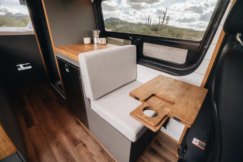 A dinette seat and fold-out wood table inside a Papago van, desert and saguaros out the window